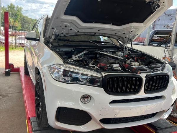 A white BMW X5 SUV on a red hydraulic lift with open hood undergoing professional auto repairs in Seffner by BMW and More Auto Care